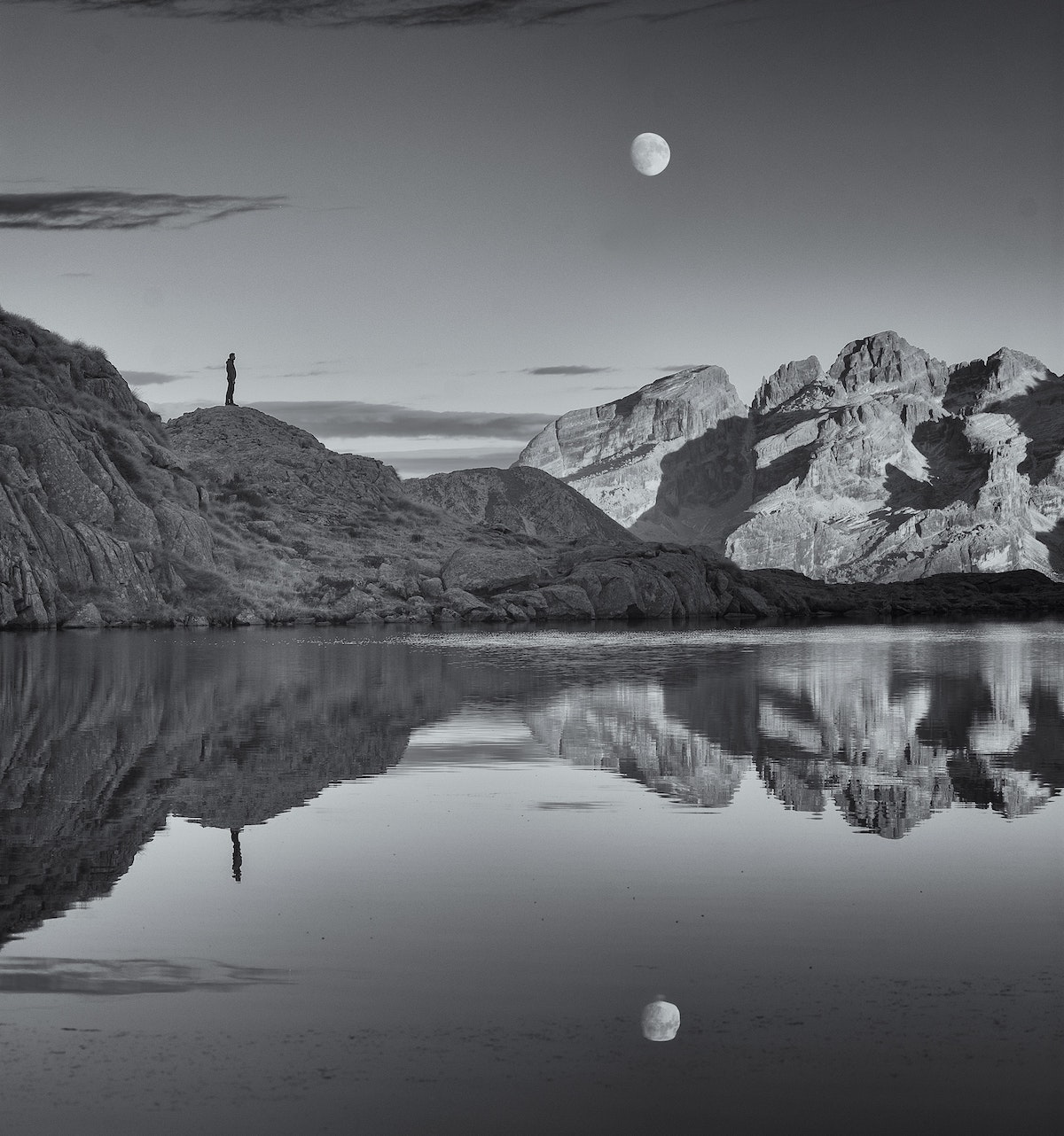 Image of a person on a rocky perch looking at a moonrise reflect on the water